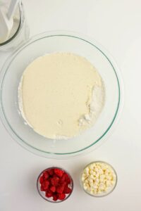 A glass bowl with pancake batter sits beside two small bowls containing raspberries and white chocolate chips, on a white surface.