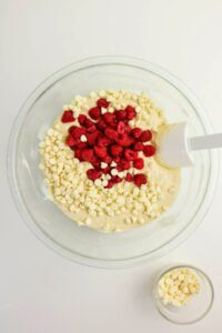 A glass mixing bowl filled with batter, white chocolate chips, and raspberries, with a spatula resting inside. A small bowl of white chocolate chips is beside it.