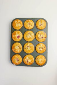 A muffin tin with twelve freshly baked muffins containing visible pieces of white chocolate and raspberries, on a white background.