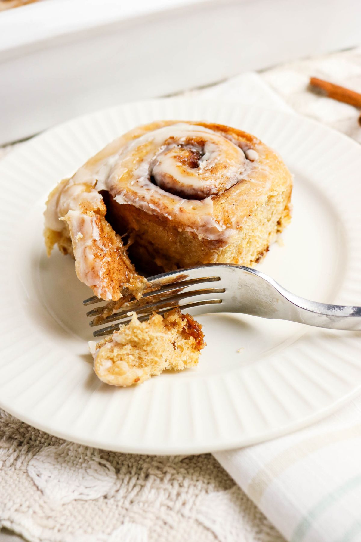 A cinnamon roll with icing on a white plate, partially cut with a fork.