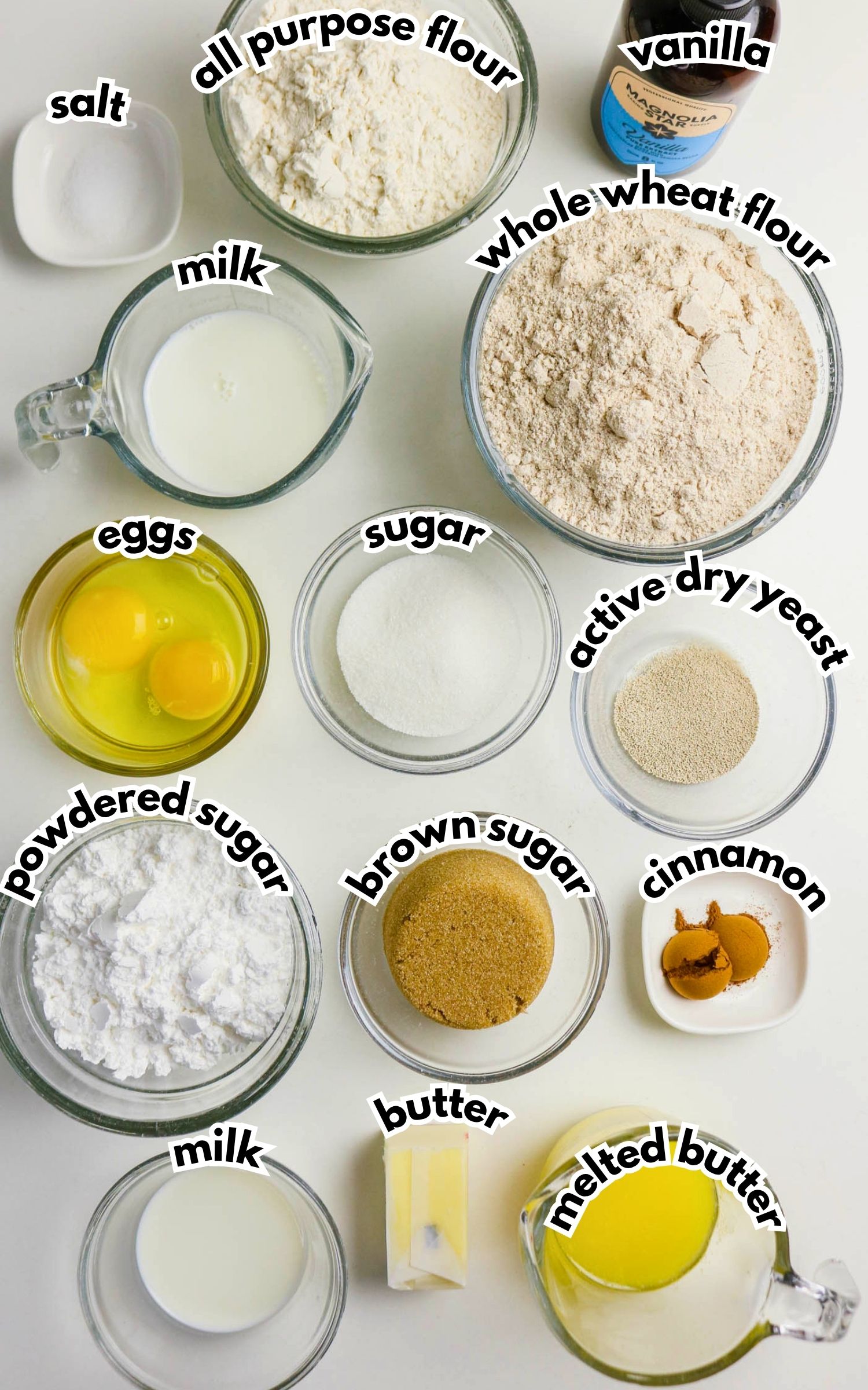 Various baking ingredients in bowls and measuring cups, including flours, sugars, eggs, milk, salt, butter, vanilla, active dry yeast, and cinnamon, are arranged on a white surface.