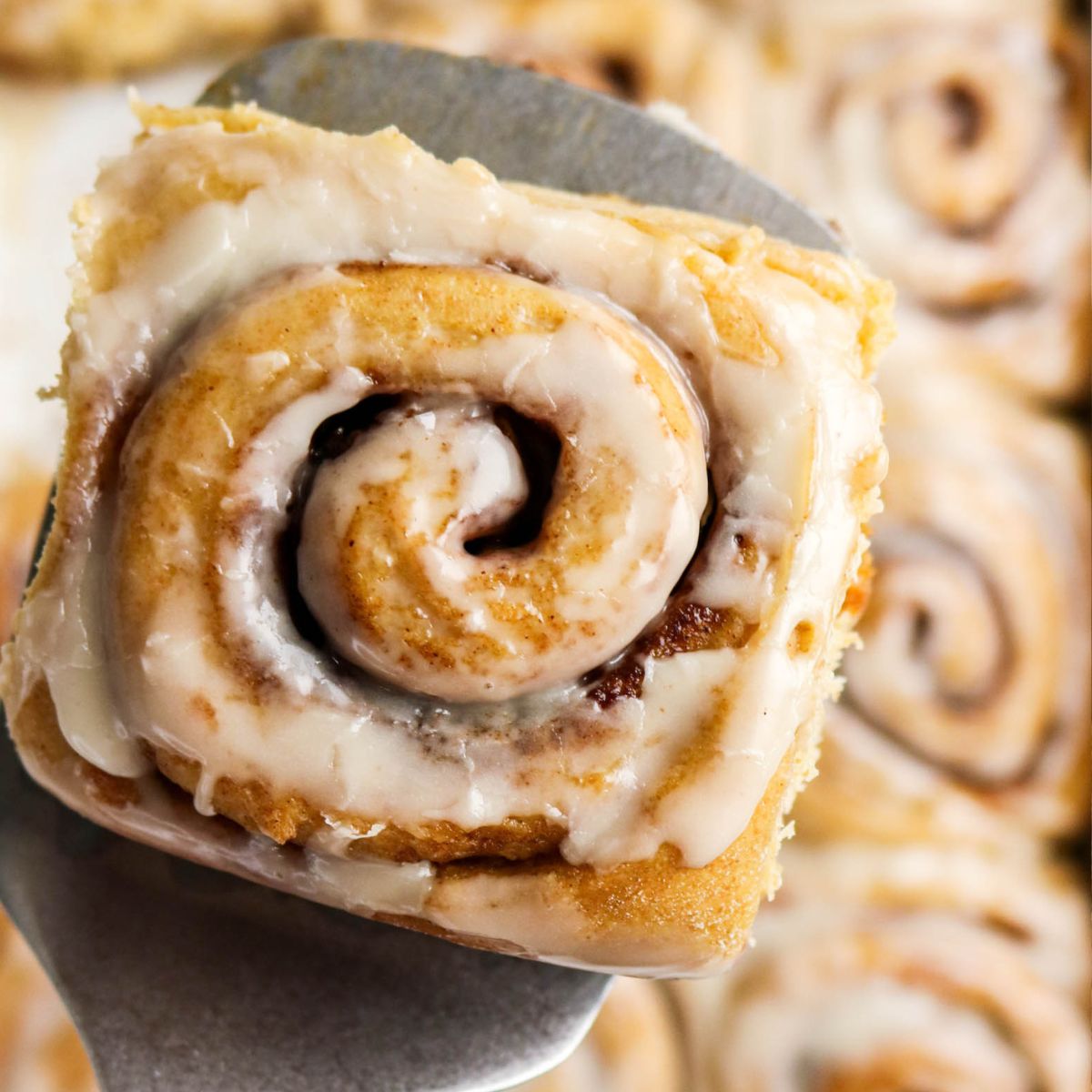 A close-up of a cinnamon roll with icing on top, held up by a metal spatula, with more cinnamon rolls visible in the background.