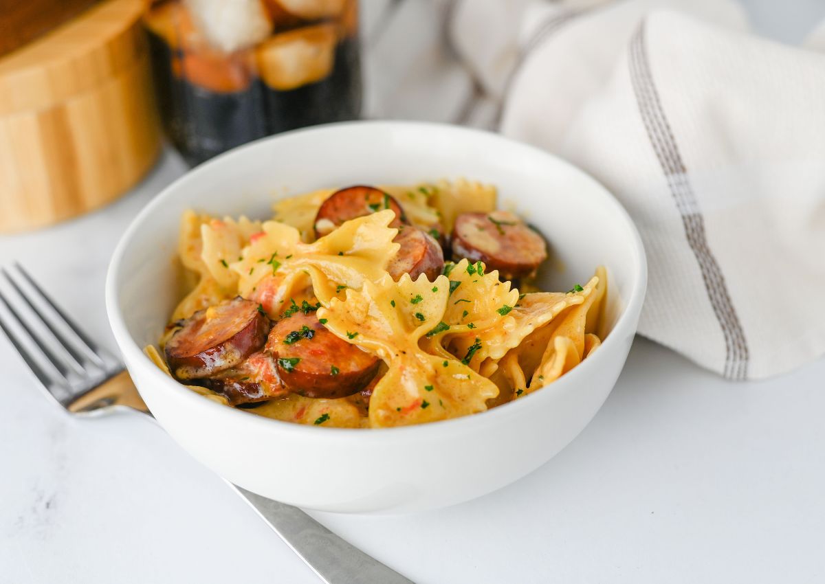 A white bowl filled with Cajun Sausage Pasta—farfalle and sliced sausage garnished with herbs—sits on a table next to a fork and a napkin.