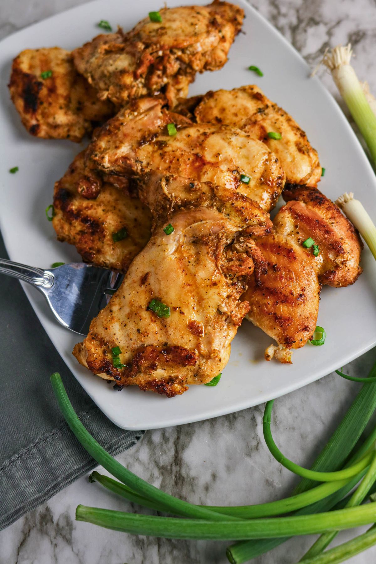 A plate of seasoned and cooked chicken breasts, complemented by Grilled Boneless Chicken Thighs and garnished with chopped green onions, with a fork and whole green onions on the side.
