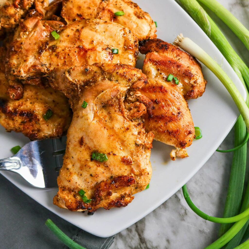 Grilled Boneless Chicken Thighs garnished with chopped green onions on a white plate, served alongside fresh green onions on a marble surface.