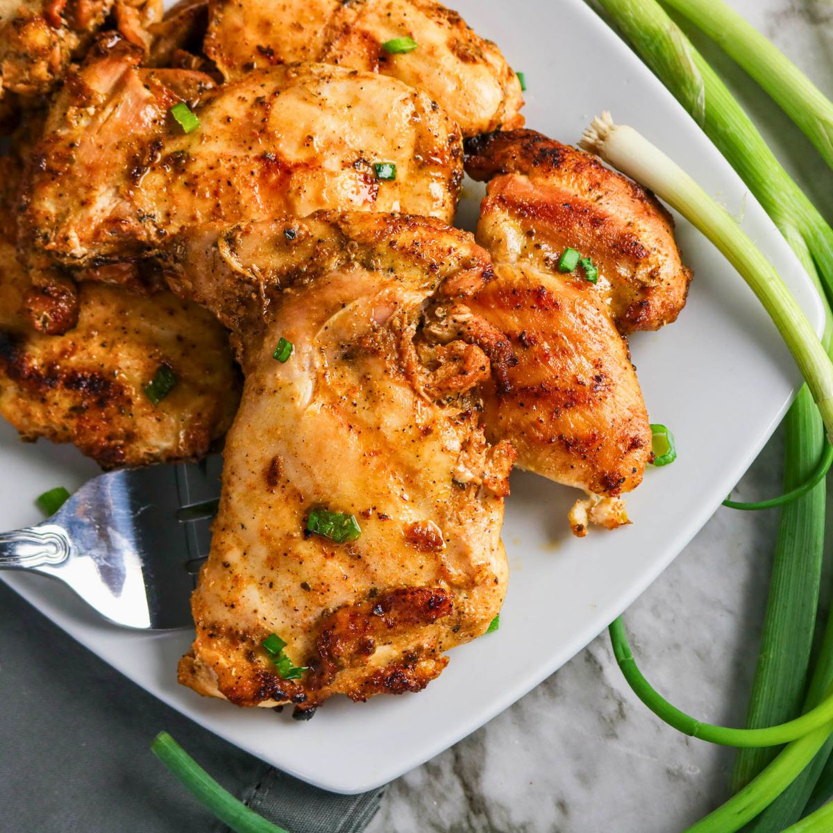 Grilled Boneless Chicken Thighs garnished with chopped green onions on a white plate, served alongside fresh green onions on a marble surface.