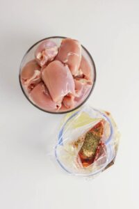 A bowl of raw chicken thighs sits beside a plastic bag filled with marinade and seasonings, ready to become tender Grilled Boneless Chicken Thighs on a white surface.