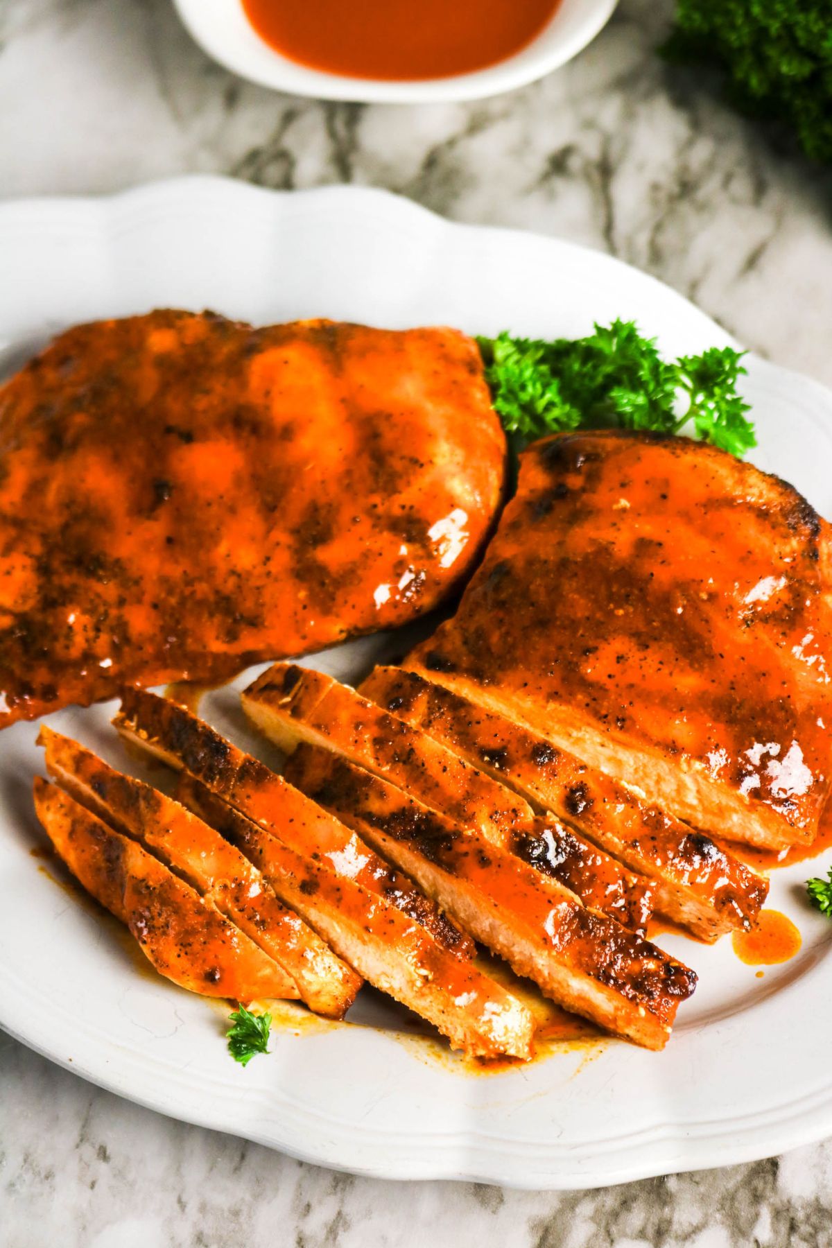 Two grilled chicken breasts coated in barbecue sauce, one whole and one sliced, served on a white plate with parsley garnish.