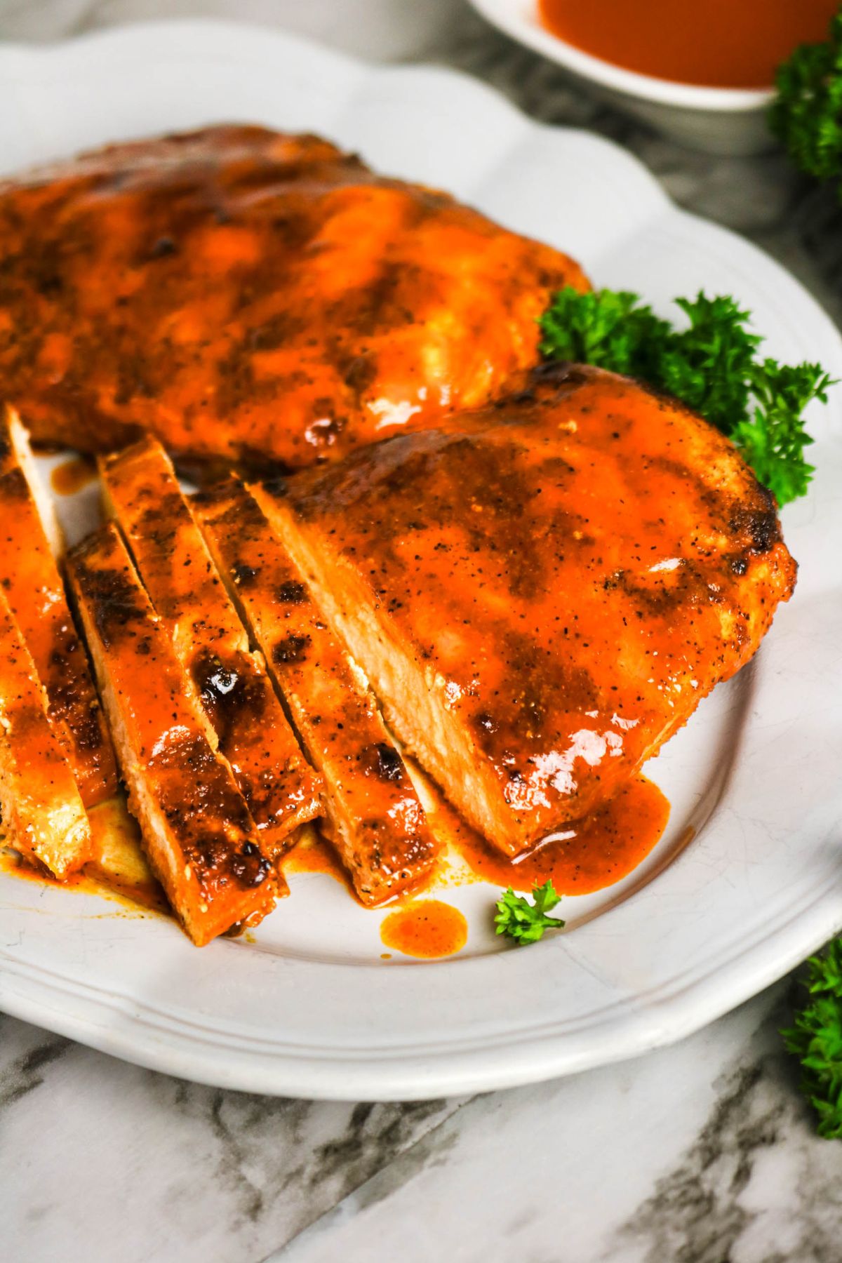 Grilled chicken breasts coated in a reddish-brown sauce, one sliced, served on a white plate with parsley garnish.