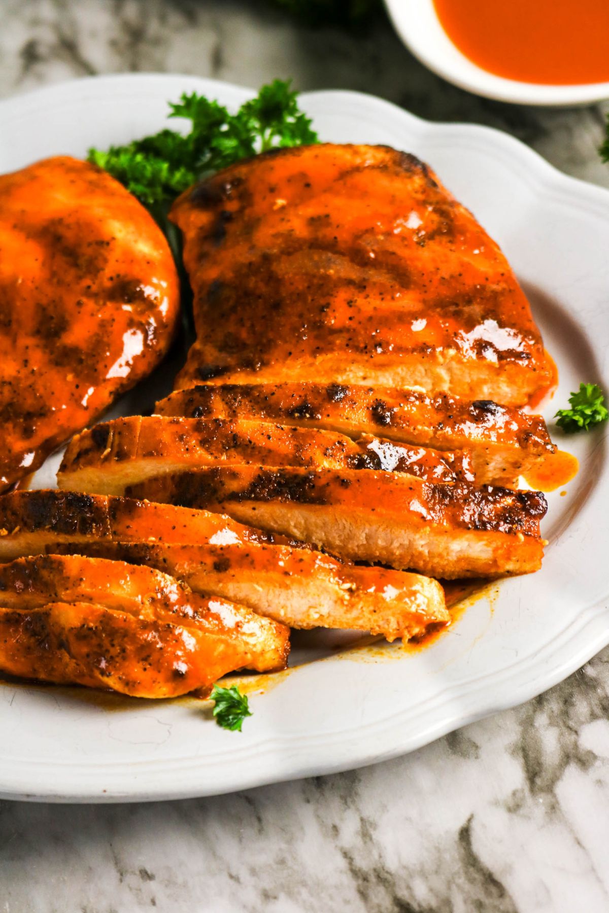 Two grilled chicken breasts coated in orange sauce on a white plate, with one breast sliced and garnished with parsley.
