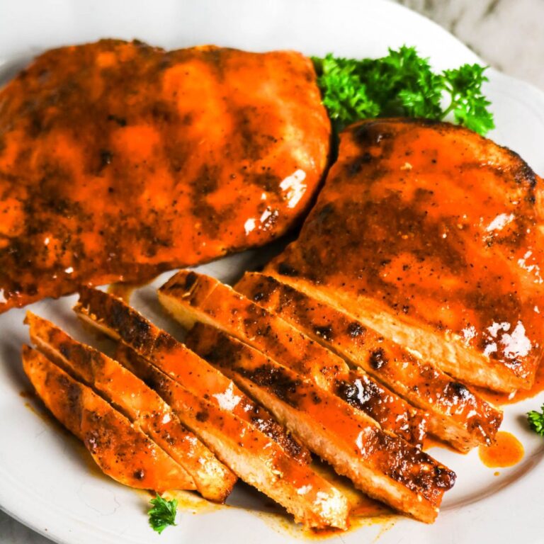 Grilled chicken breasts covered in barbecue sauce, with one piece sliced, served on a white plate with parsley garnish.