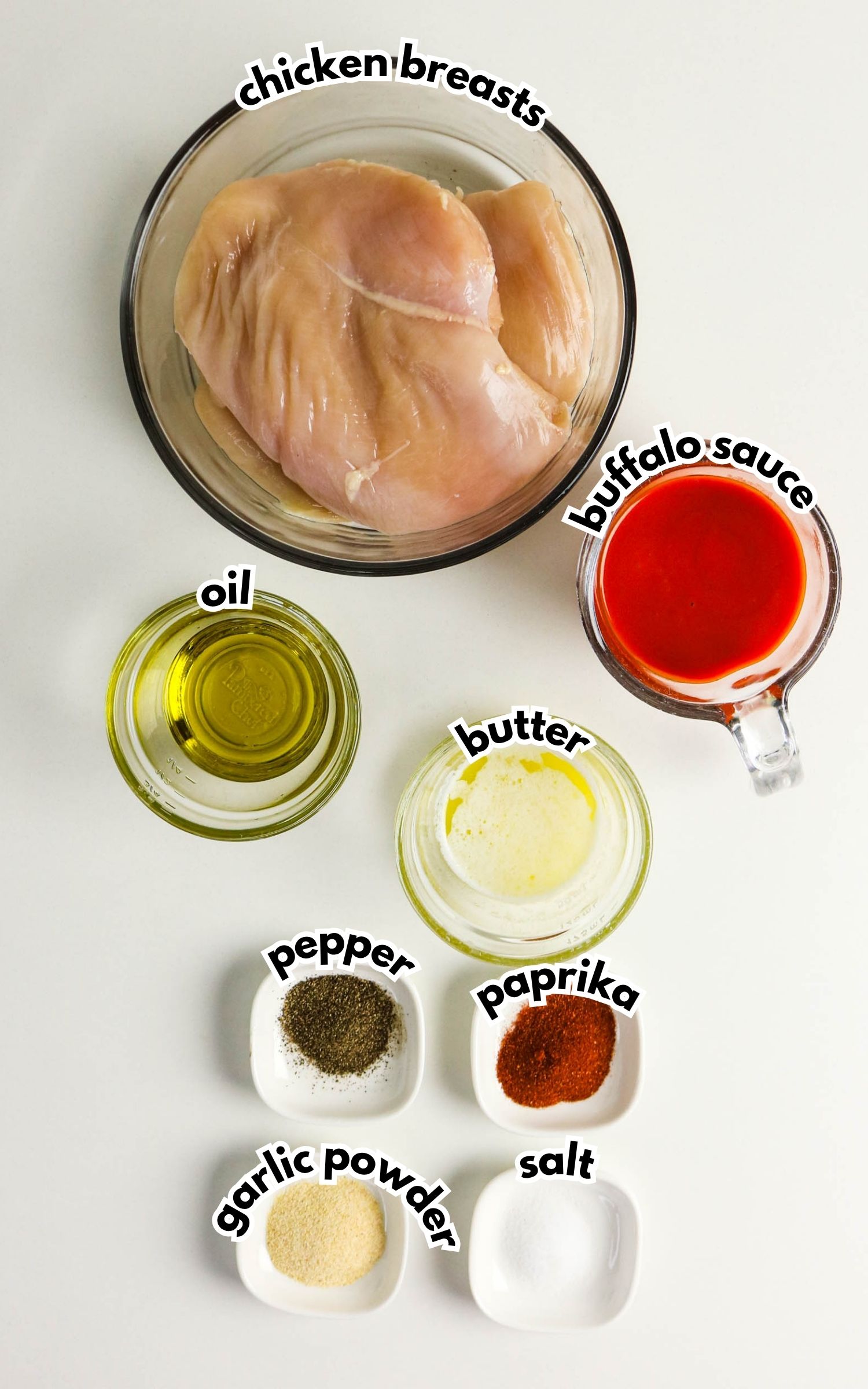 Top-down view of raw chicken breasts, buffalo sauce, oil, butter, pepper, paprika, garlic powder, and salt in separate bowls on a white background, each labeled with text.