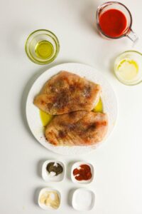 Two raw chicken breasts seasoned on a white plate, surrounded by small bowls containing olive oil, melted butter, buffalo sauce, and various spices on a white surface.