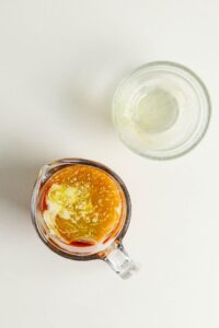 A glass measuring cup with oil, vinegar, and seasonings next to an empty glass bowl on a white surface.