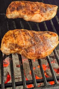 Two seasoned chicken breasts cooking on a grill over hot charcoal.