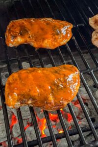 Two pieces of barbecue sauce-coated chicken are grilling over hot charcoal on a barbecue grill.