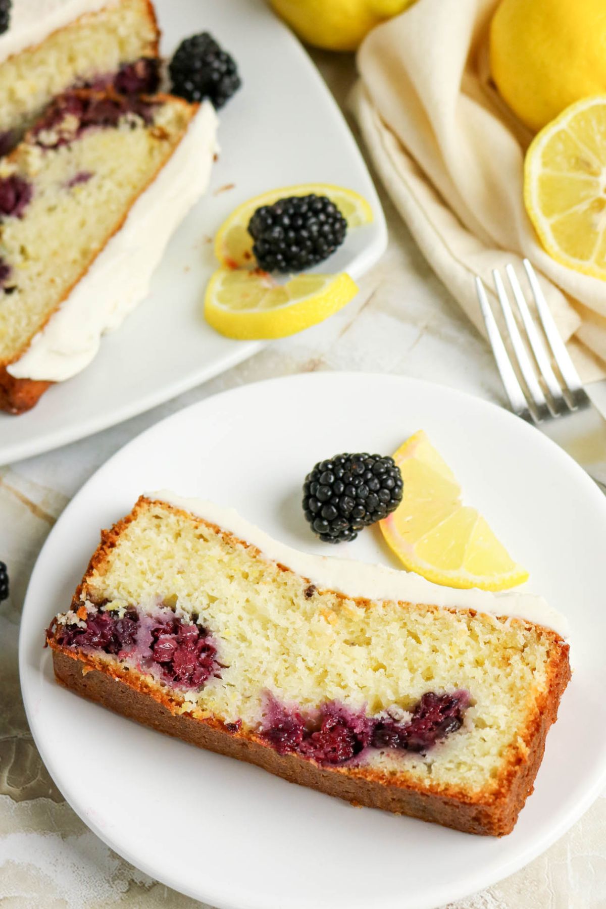 A slice of lemon blackberry loaf cake with blackberries and lemon slices on a white plate; more cake, fruit, and lemons in the background.