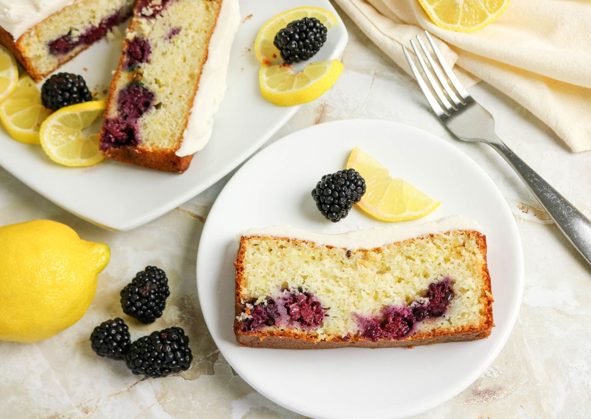 A slice of blackberry lemon loaf cake on a white plate, garnished with lemon wedges and blackberries, with more cake and fruit in the background.