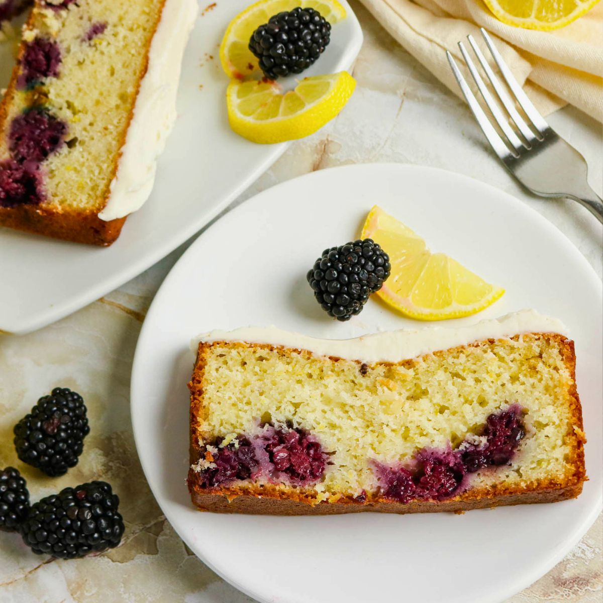 A slice of blackberry lemon loaf cake with icing is served on a white plate, garnished with a blackberry and lemon wedges; more cake and fruit are nearby.