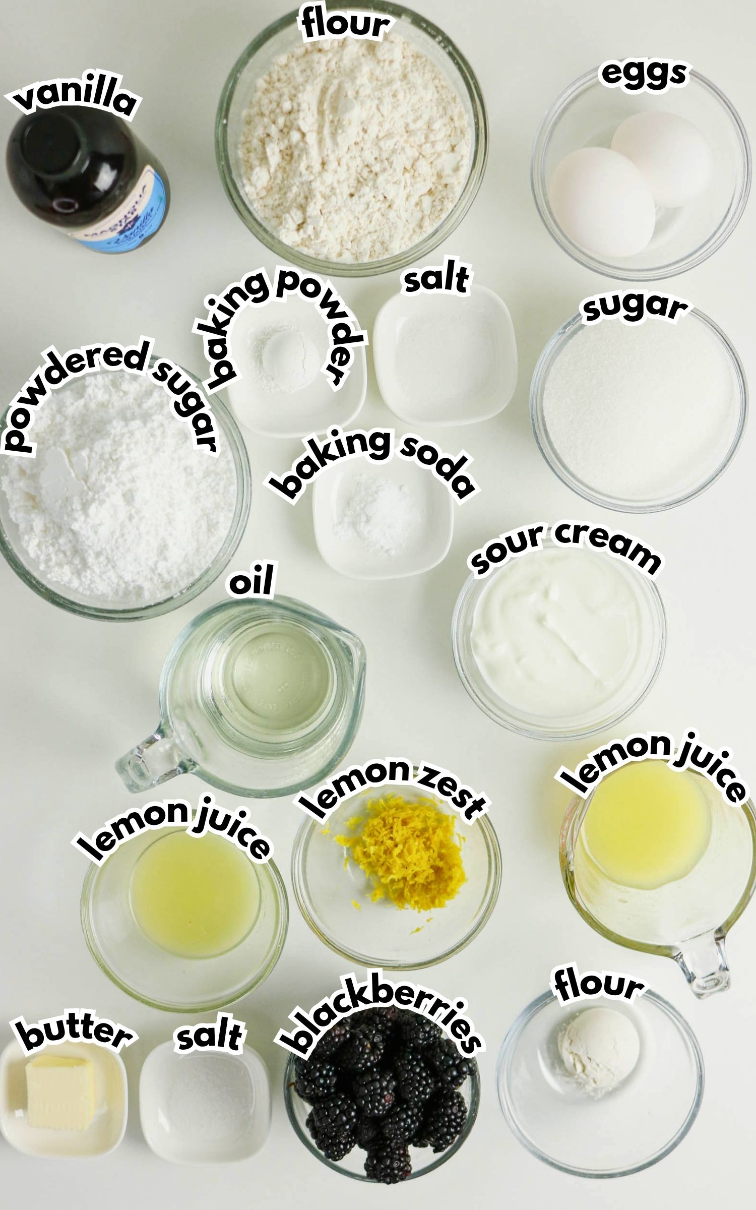 Various baking ingredients in bowls, including flour, eggs, sugar, vanilla, powdered sugar, baking powder, baking soda, sour cream, lemon zest, lemon juice, butter, salt, and blackberries.