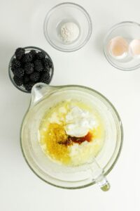 A mixing bowl with wet ingredients and three small bowls containing blackberries, flour, and eggshells on a white surface.