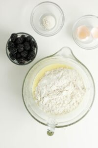 Glass mixing bowl with flour and batter, next to small bowls of blackberries, baking powder, and two eggshell halves on a white surface.