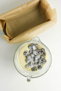 A glass mixing bowl with cake batter and floured blackberries next to a baking pan lined with parchment paper on a white surface.