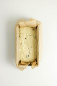 A loaf pan lined with parchment paper filled with unbaked batter containing visible chocolate chips or fruit pieces, viewed from above.