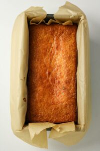 A loaf of golden-brown bread sits in a rectangular baking pan lined with parchment paper on a white surface.