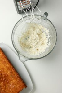 A glass mixing bowl with whipped frosting and electric mixer attachments next to a partially visible rectangular baked cake on a white surface.