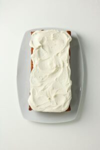 A rectangular loaf cake with white frosting is placed on a white rectangular plate against a plain white background.