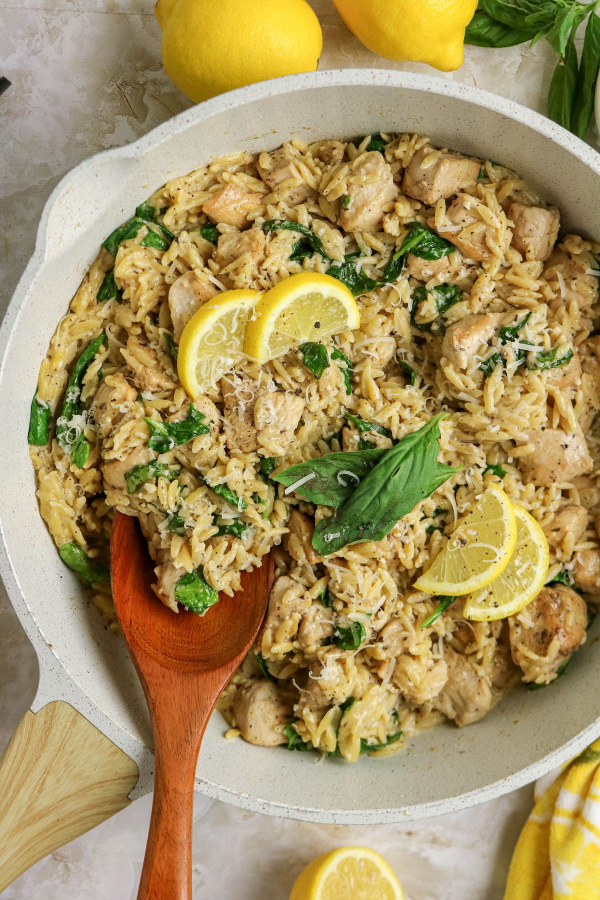 A skillet filled with creamy orzo, cooked chicken pieces, spinach, and garnished with lemon slices and basil leaves, with a wooden spoon resting inside.