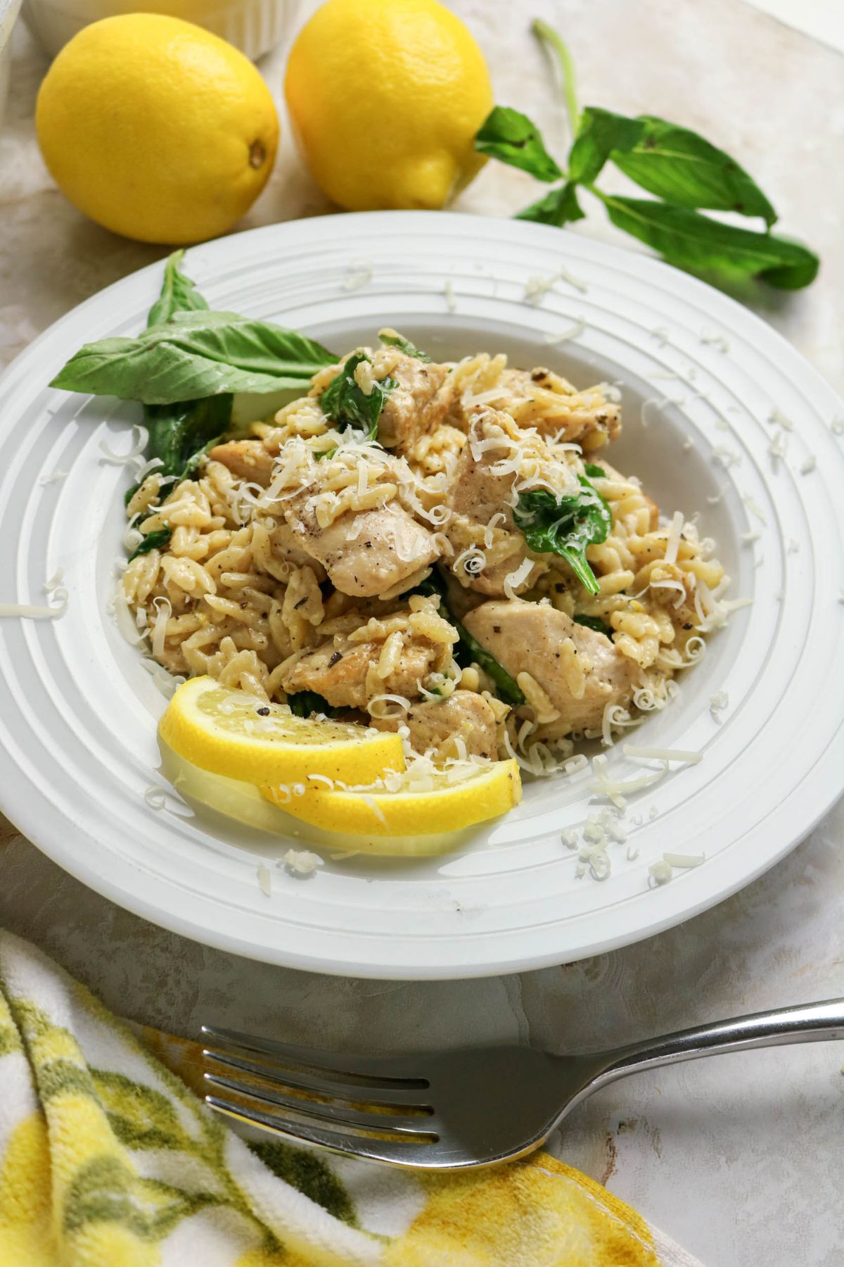 A white plate of chicken pasta with spinach, topped with shredded cheese and a lemon wedge, sits on a table next to fresh lemons, basil leaves, and a fork.