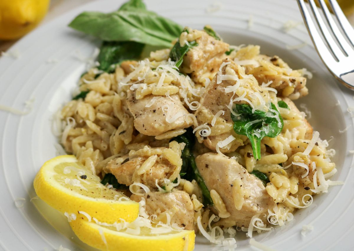 A plate of orzo pasta with pieces of chicken, spinach, grated cheese, and lemon slices, garnished with fresh basil.