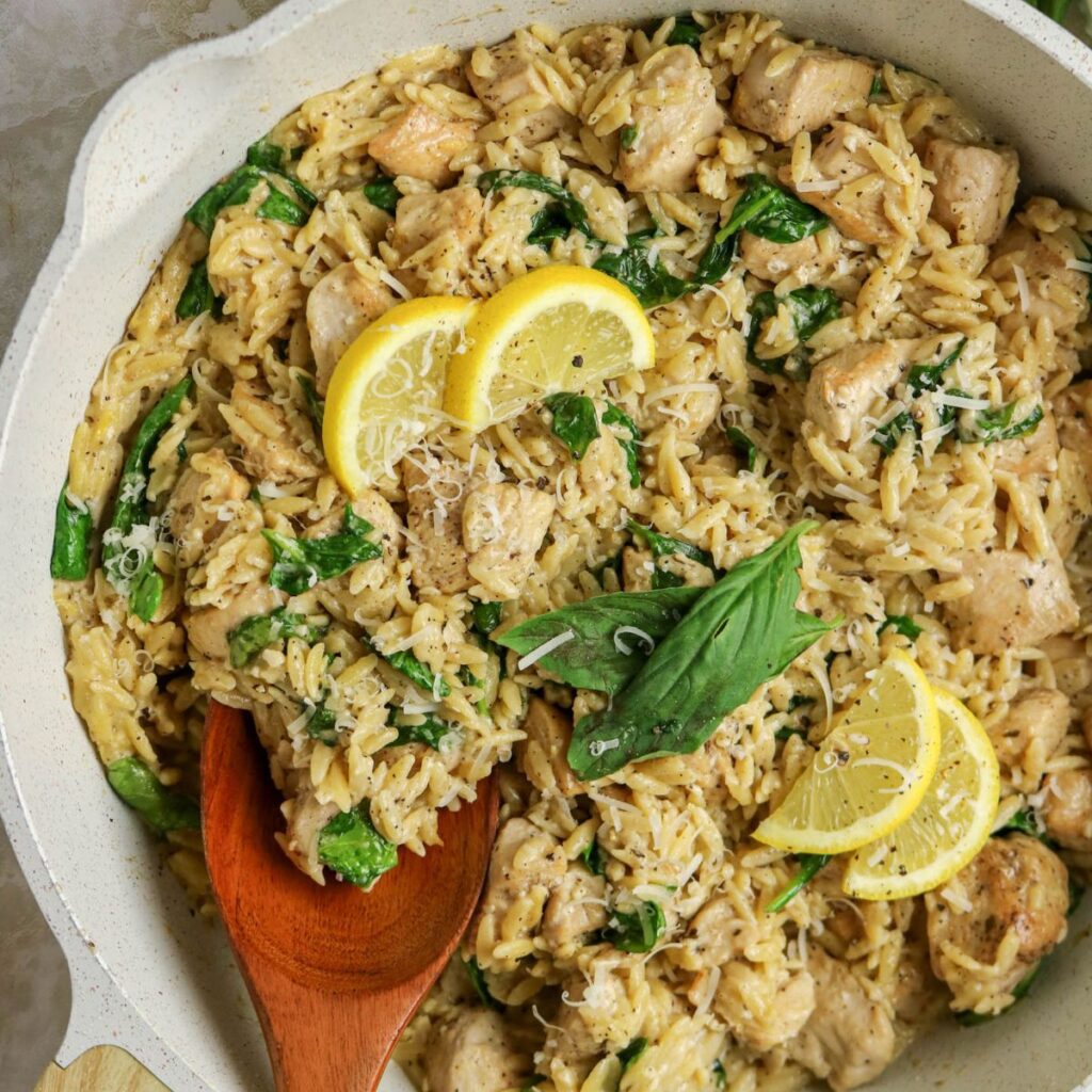A skillet filled with creamy orzo pasta, chicken pieces, spinach, grated cheese, and garnished with basil leaves and lemon slices. A wooden spoon rests in the pan.