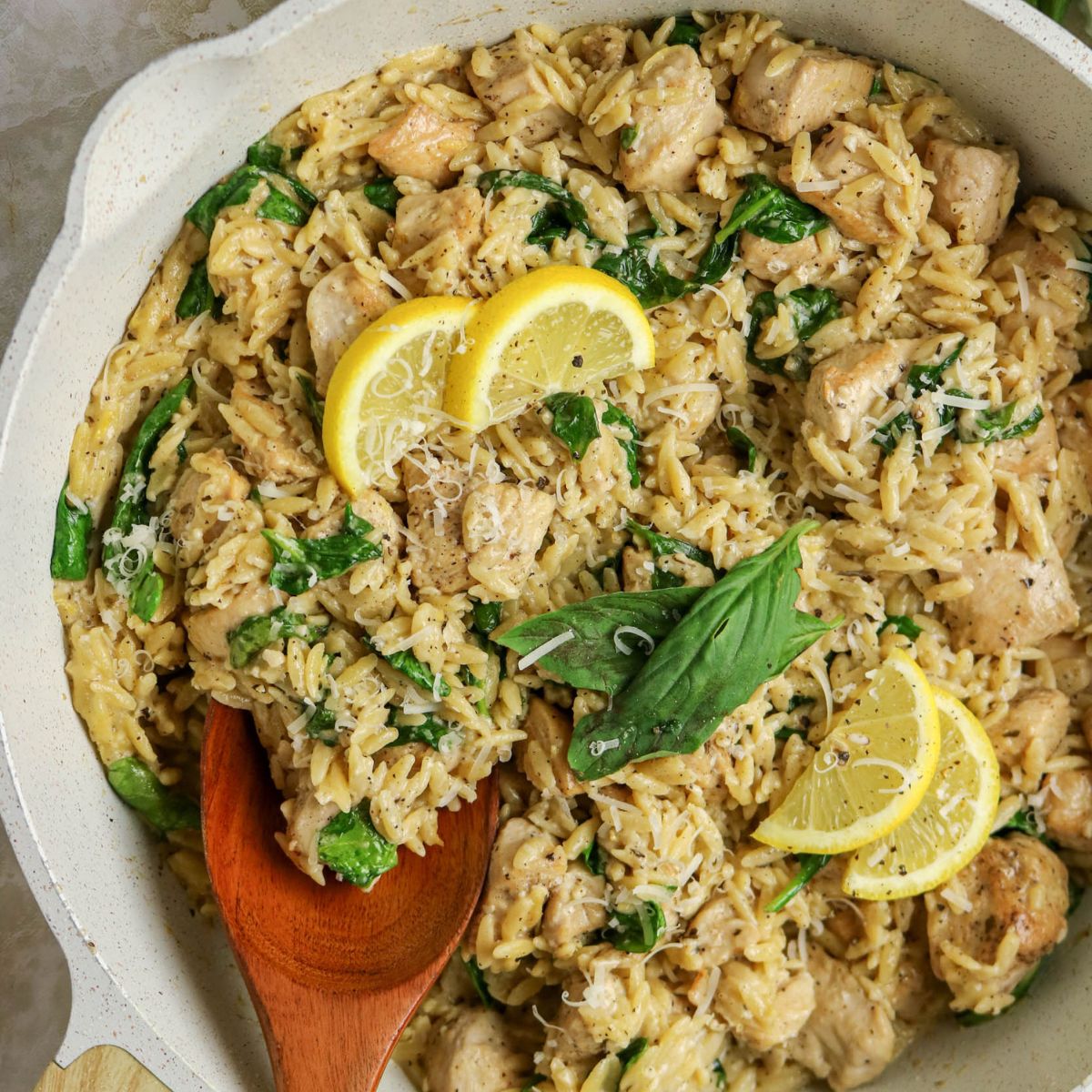A skillet filled with creamy orzo pasta, chicken pieces, spinach, grated cheese, and garnished with basil leaves and lemon slices. A wooden spoon rests in the pan.