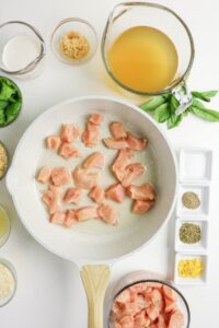 Raw chicken pieces are cooking in a white skillet, surrounded by small bowls containing broth, cream, herbs, garlic, cheese, and other ingredients on a white surface.