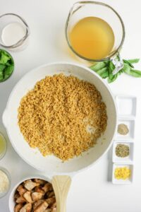 Overhead view of a skillet with toasted rice, surrounded by bowls of broth, cream, spices, fresh basil, parmesan, lemon zest, and cooked sausage on a white surface.