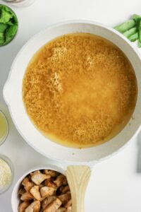 A white pan on a stove contains uncooked rice in broth. Nearby are bowls with cooked chicken, grated cheese, fresh basil, and other ingredients.