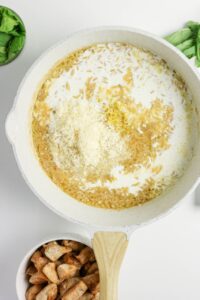 A white pan with orzo, milk, and shredded cheese is on a white surface; bowls of spinach and cooked chicken are nearby.