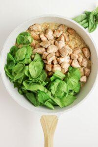 A skillet with cooked diced chicken, fresh spinach leaves, and creamy rice, viewed from above.