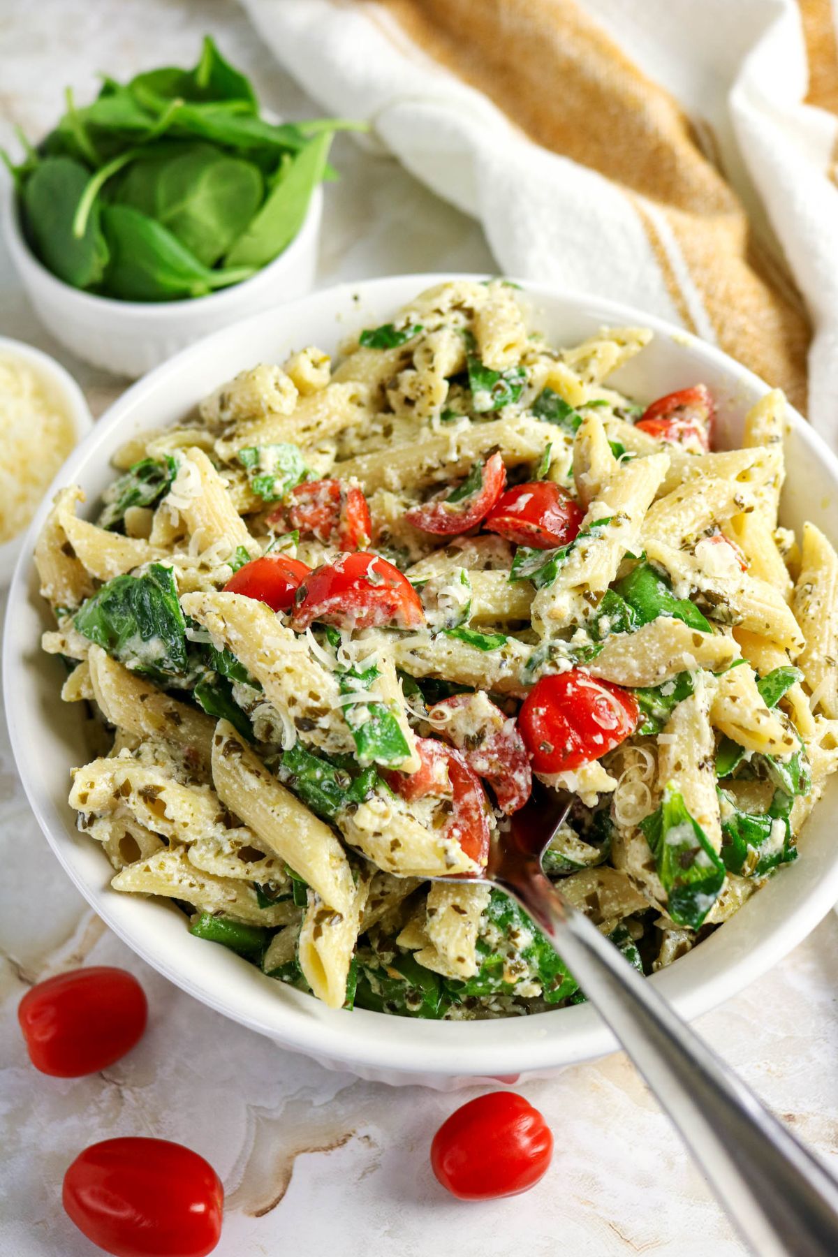A bowl of penne pasta mixed with spinach, cherry tomatoes, grated cheese, and a creamy sauce. A spoon rests in the bowl, with extra spinach and tomatoes nearby.