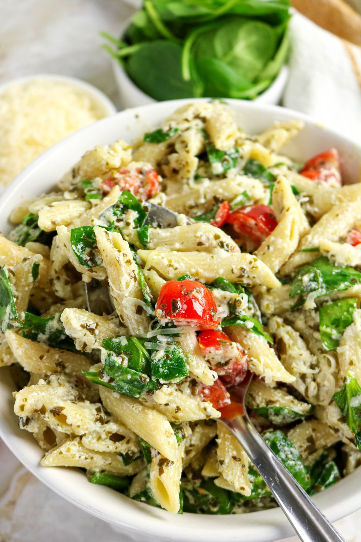 A bowl of penne pasta mixed with spinach, cherry tomatoes, and grated cheese, with a spoon inside. A small bowl of spinach and a container of grated cheese are in the background.