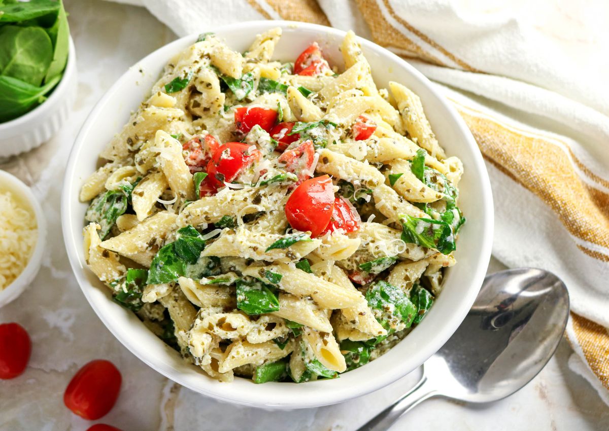 A white bowl filled with penne pasta mixed with pesto, spinach, cherry tomatoes, and shredded cheese, placed on a white surface with a spoon beside it.