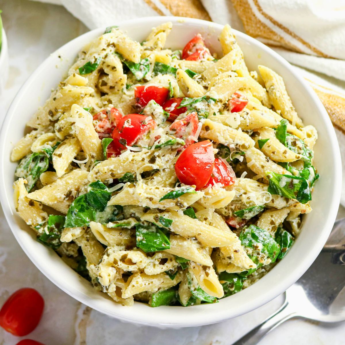 A bowl of penne pasta mixed with spinach, cherry tomatoes, grated cheese, and a creamy sauce, garnished with extra tomatoes.
