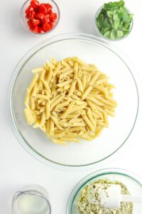 A glass bowl of cooked penne pasta is on a white surface, surrounded by bowls of cherry tomatoes, fresh basil, milk, and a creamy herb sauce.