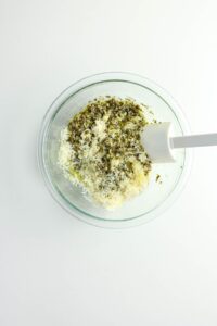 A glass bowl containing shredded cheese, pesto, and a white spatula on a white surface.