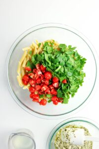 A glass bowl contains uncooked penne pasta, chopped spinach, and halved cherry tomatoes. Nearby are a measuring cup with liquid and a bowl with a white and green sauce.