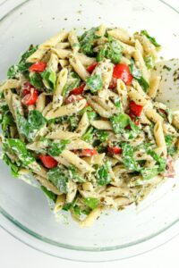 Penne pasta mixed with spinach, cherry tomatoes, pesto, and grated cheese in a clear glass bowl with a white spoon.