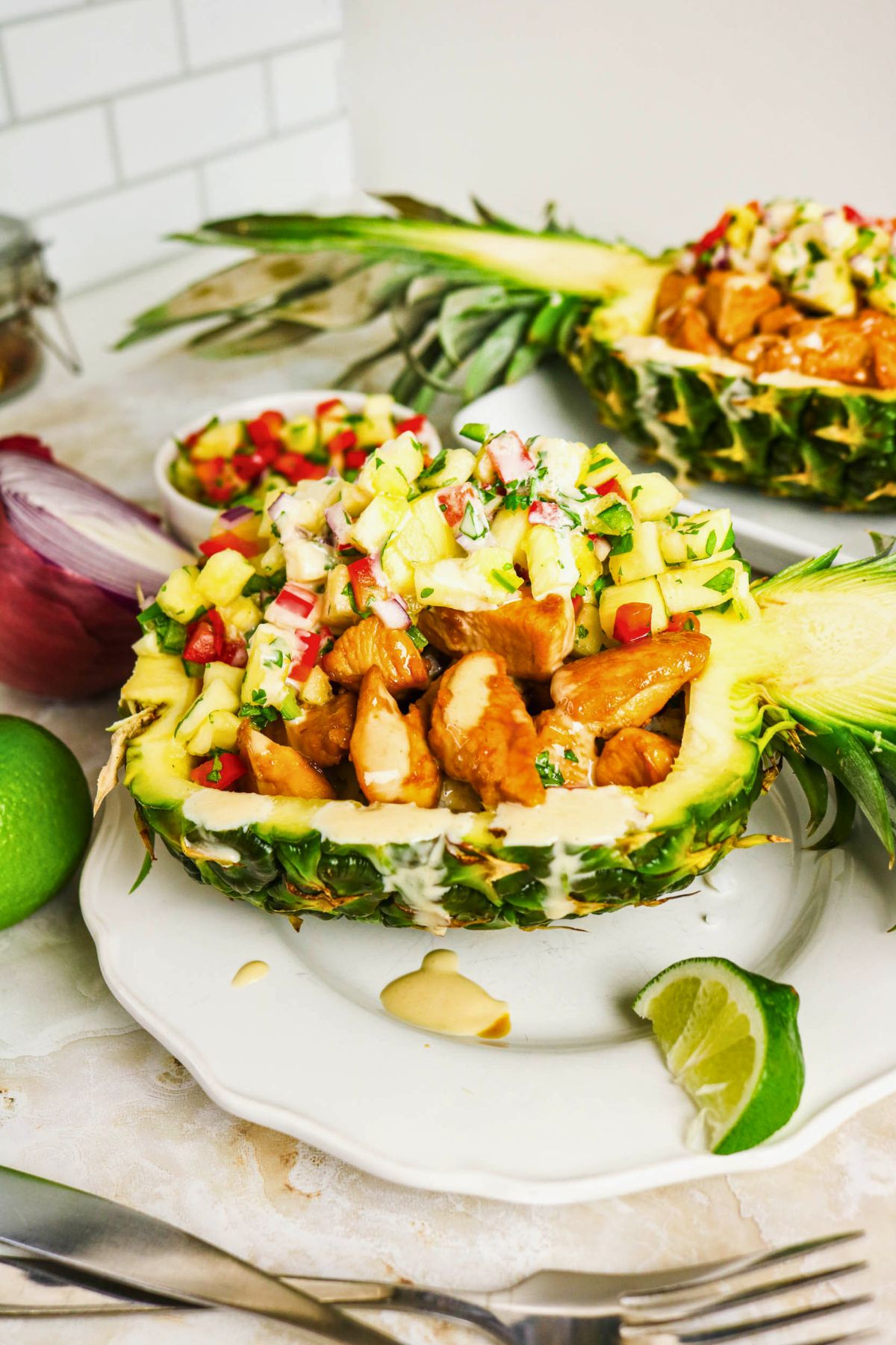 Pineapple Chicken Bowls feature a pineapple half used as a bowl, filled with tender cooked chicken and topped with zesty pineapple salsa, served on a white plate with fresh lime wedges.